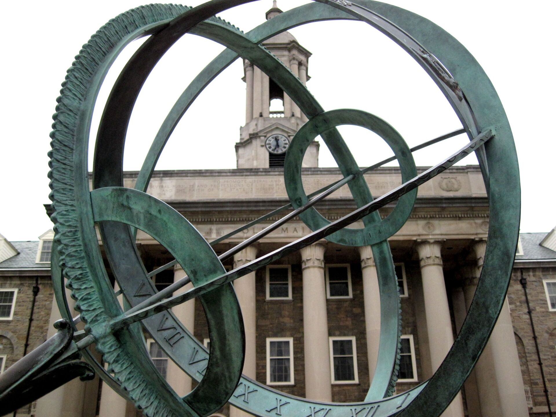 Bronze with patina orbit sculpture in focus with Old Main in background, white sky.