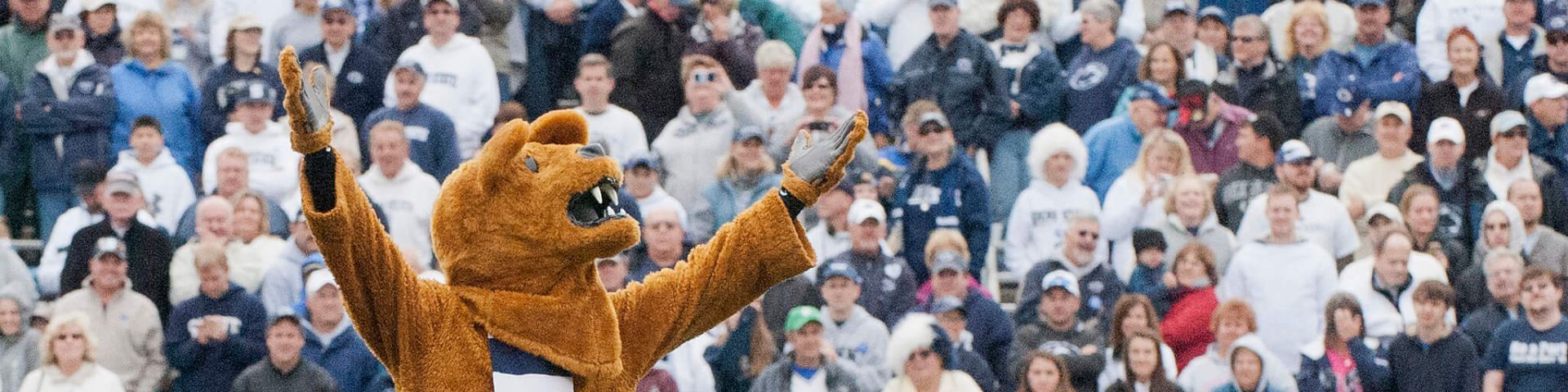 Nittany Lion mascot standing on football turf with arms raised in front of Penn State football fans mainly wearing white and blue.