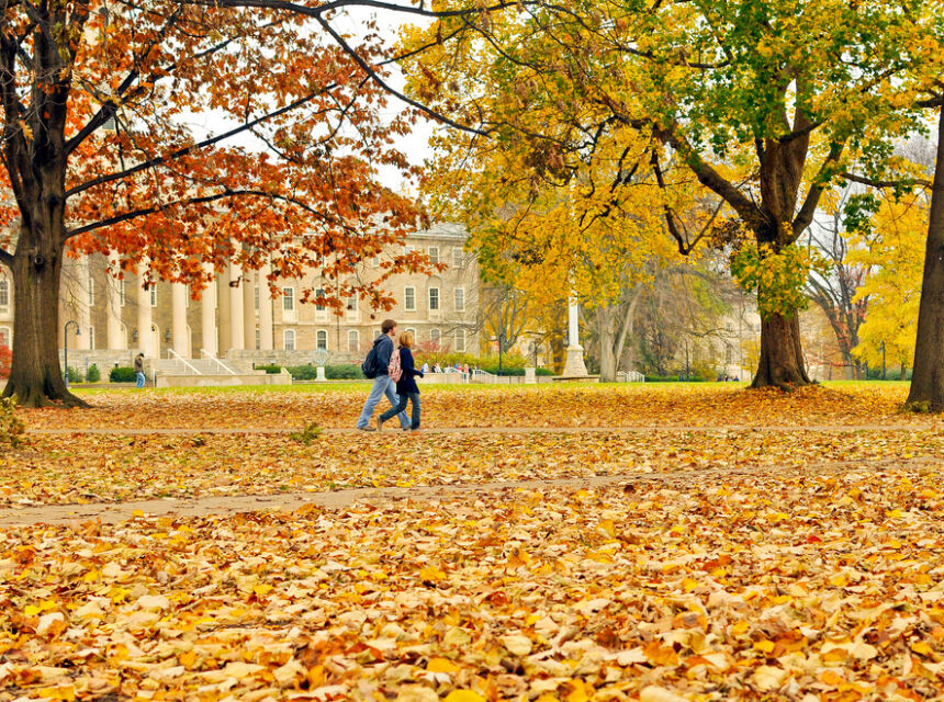 Grounds in front of Old Main covered in golden leaves. Trees with red and yellow leaves. Two people walking on path mid-ground. Columns of Old Main visible in distance.